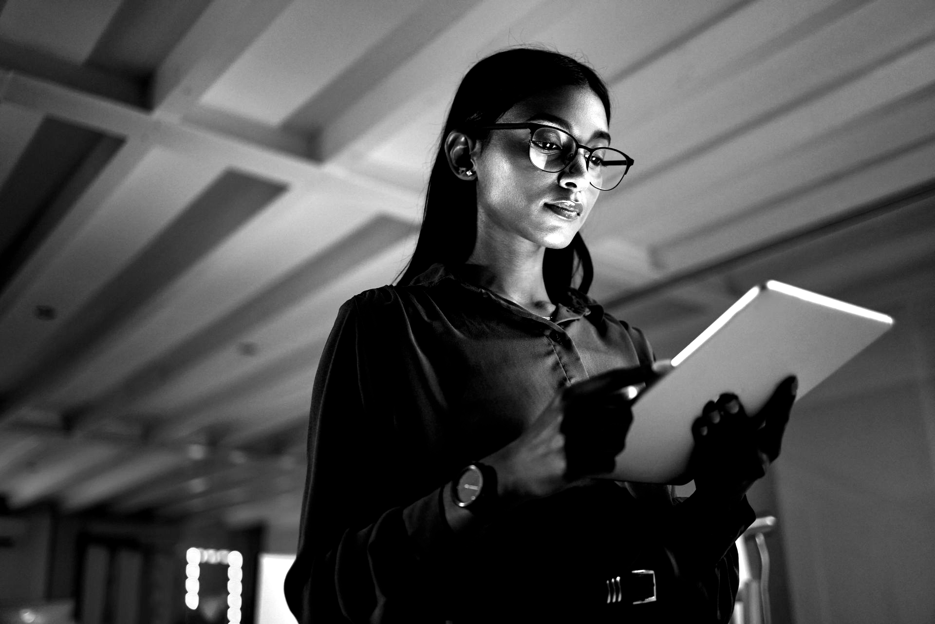 A professional wearing glasses holds a tablet in an office space, looking at the screen with a focused expression.