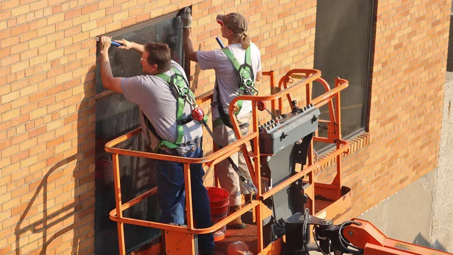 Two construction workers in an orange machine holding them up to a building so they can install windows. Two construction workers in an orange machine holding them up to a building so they can install windows.