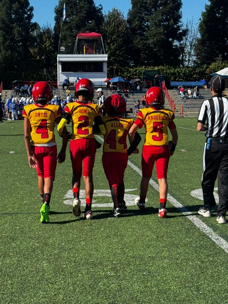 Youth football players in red and yellow uniforms walk onto the field with a referee nearby.