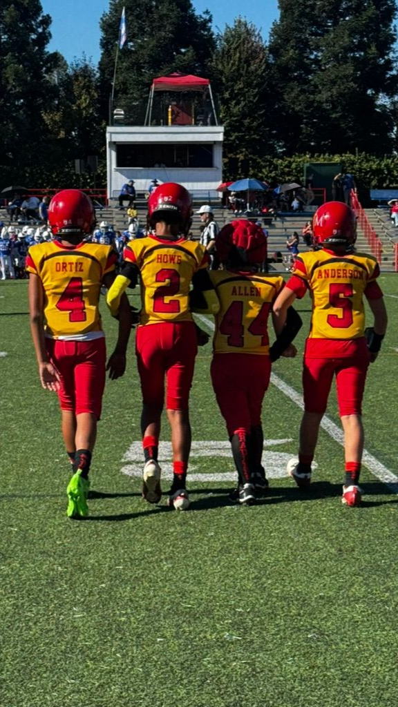 Four youth football players in red helmets and yellow jerseys walk onto a green field.