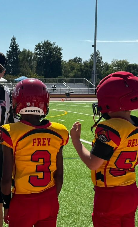 Youth football players in red-and-yellow uniforms watch from the sideline on a sunny field.