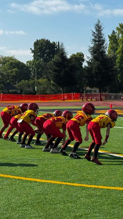 Youth football team crouched at the line on a sunny field, wearing red and yellow uniforms.