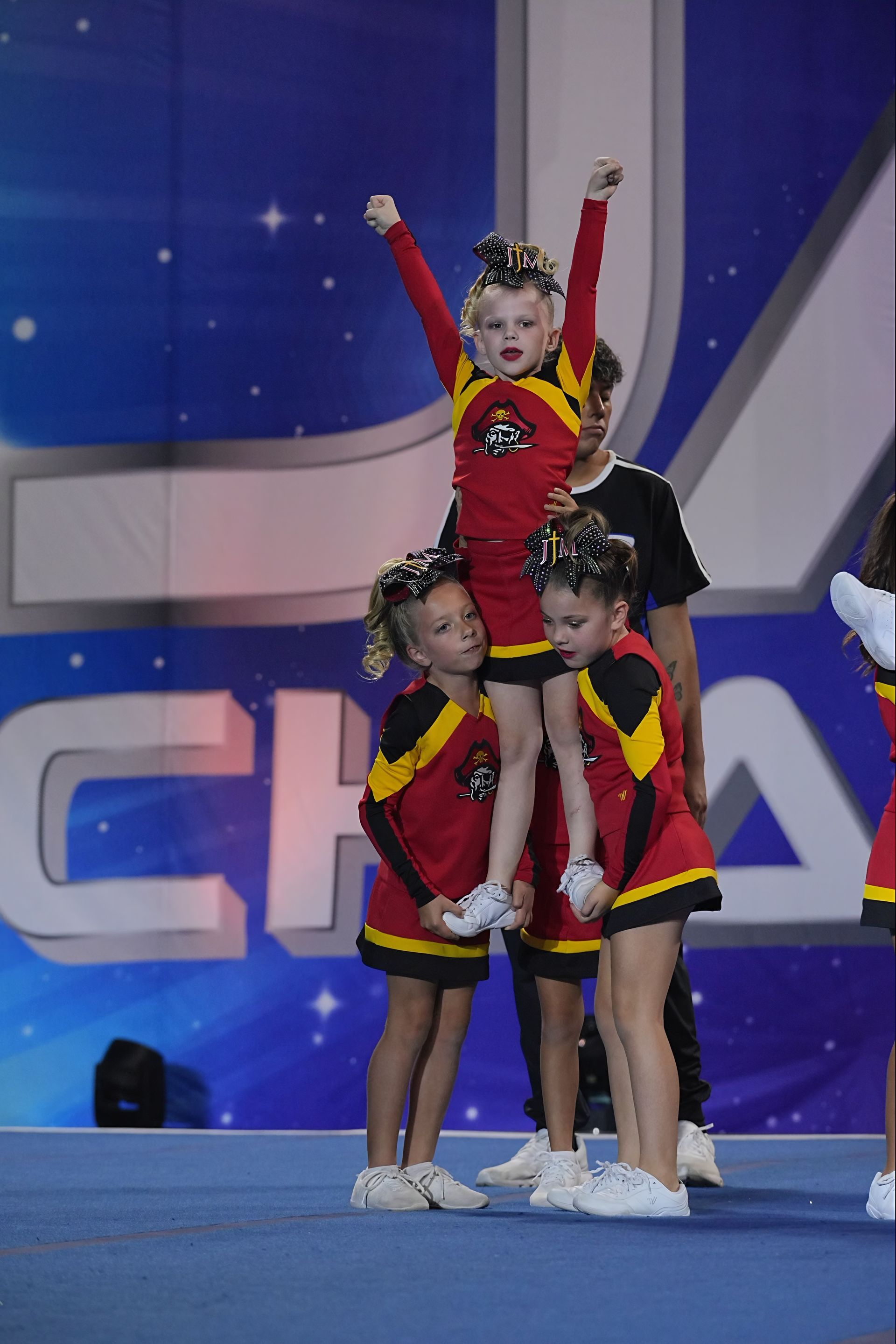 Three cheerleaders in red and black uniforms posing on a blue stage, one raised with arms up.