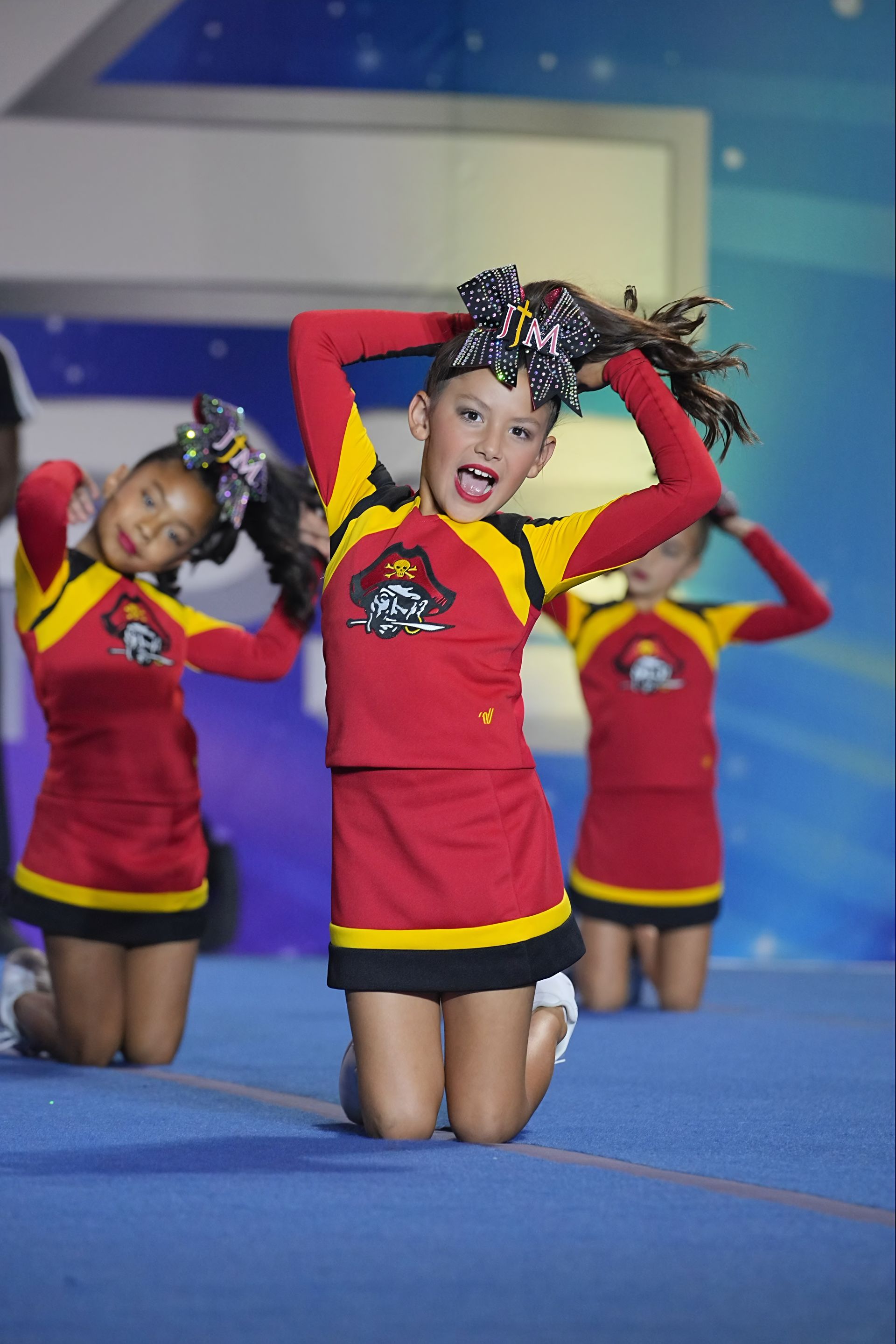 Three cheerleaders in red uniforms kneel on a blue stage, posing with hands on their heads.