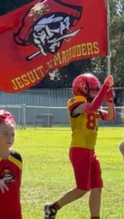 Youth football player in red and gold raising a Jesuit Marauders flag on a sunny field