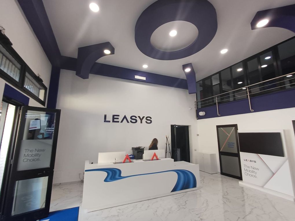 Area reception moderna nei colori bianco e blu con scrivania, logo LEASYS e porte d'ingresso in vetro.