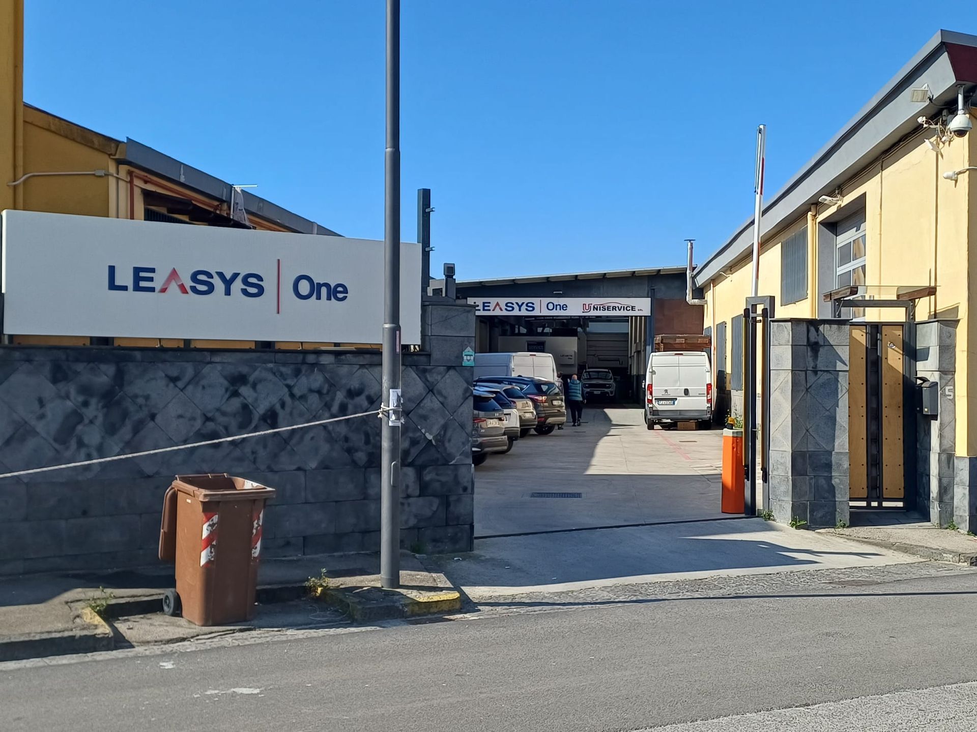 Ingresso di un magazzino industriale con insegna LEASYS, veicoli parcheggiati e vialetto recintato sotto un cielo azzurro.