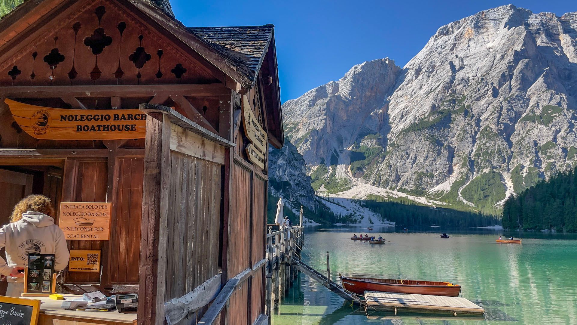 Eine Frau steht vor einem Holzgebäude neben einem See mit Bergen im Hintergrund.