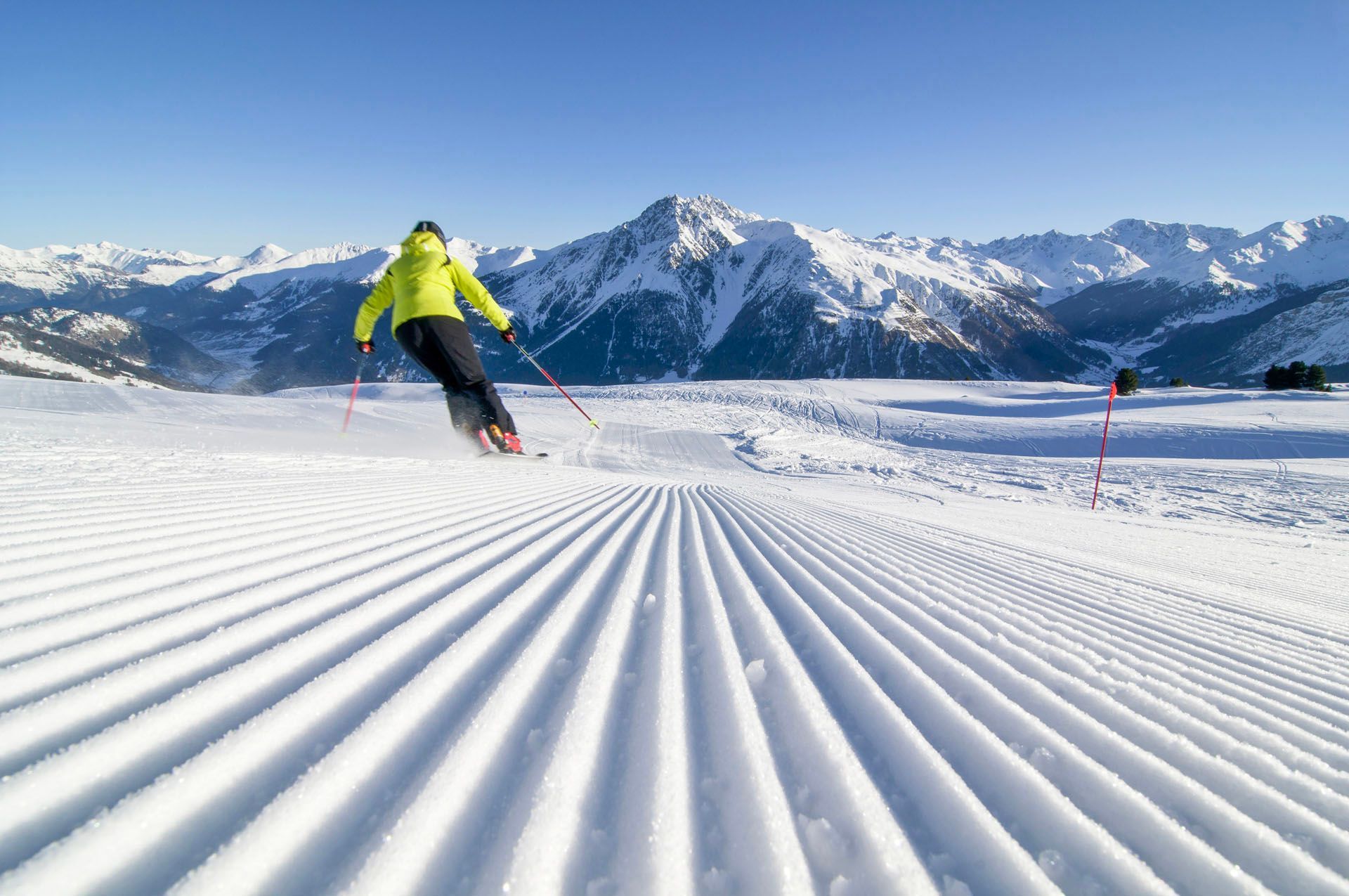 Eine Person fährt mit Skiern einen schneebedeckten Hang hinunter, im Hintergrund sind Berge zu sehen.