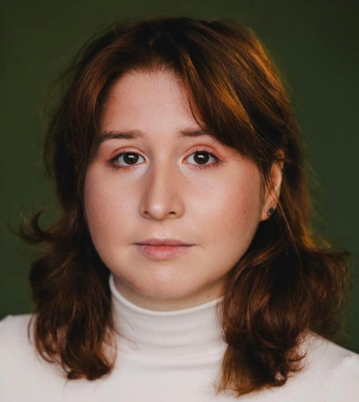 Close up photo of Rachel, she wears a white turtle neck and has browny red hair. The background is dark green. 