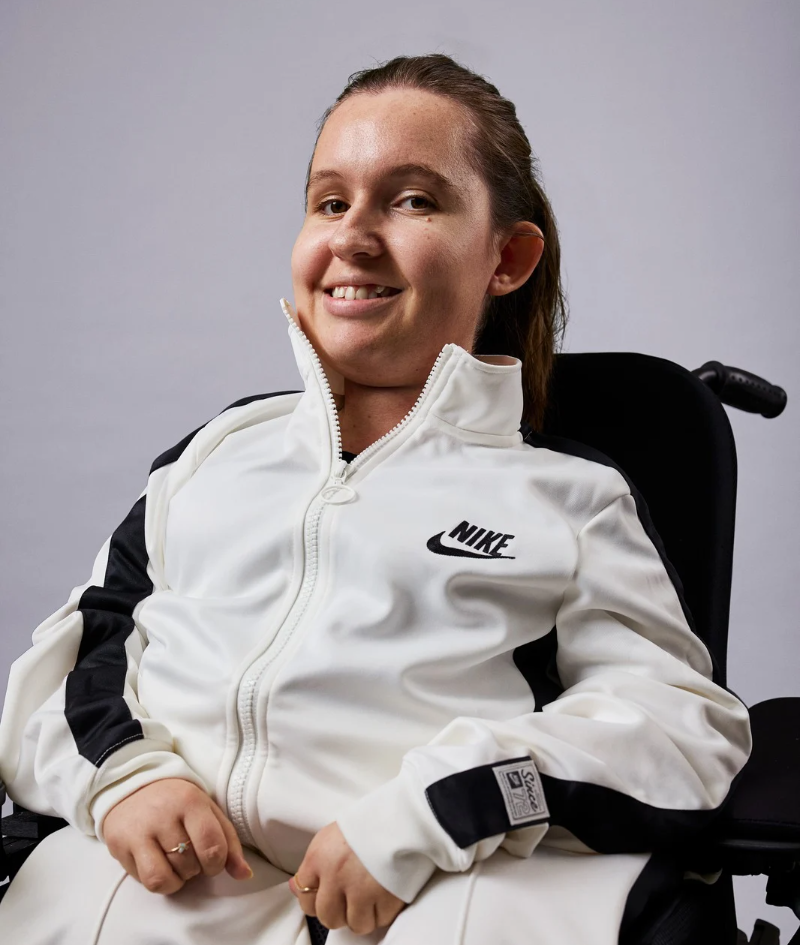 Close up studio portrait of Jamieson in her electric wheelchair, wearing a white Nike matching tracksuit.