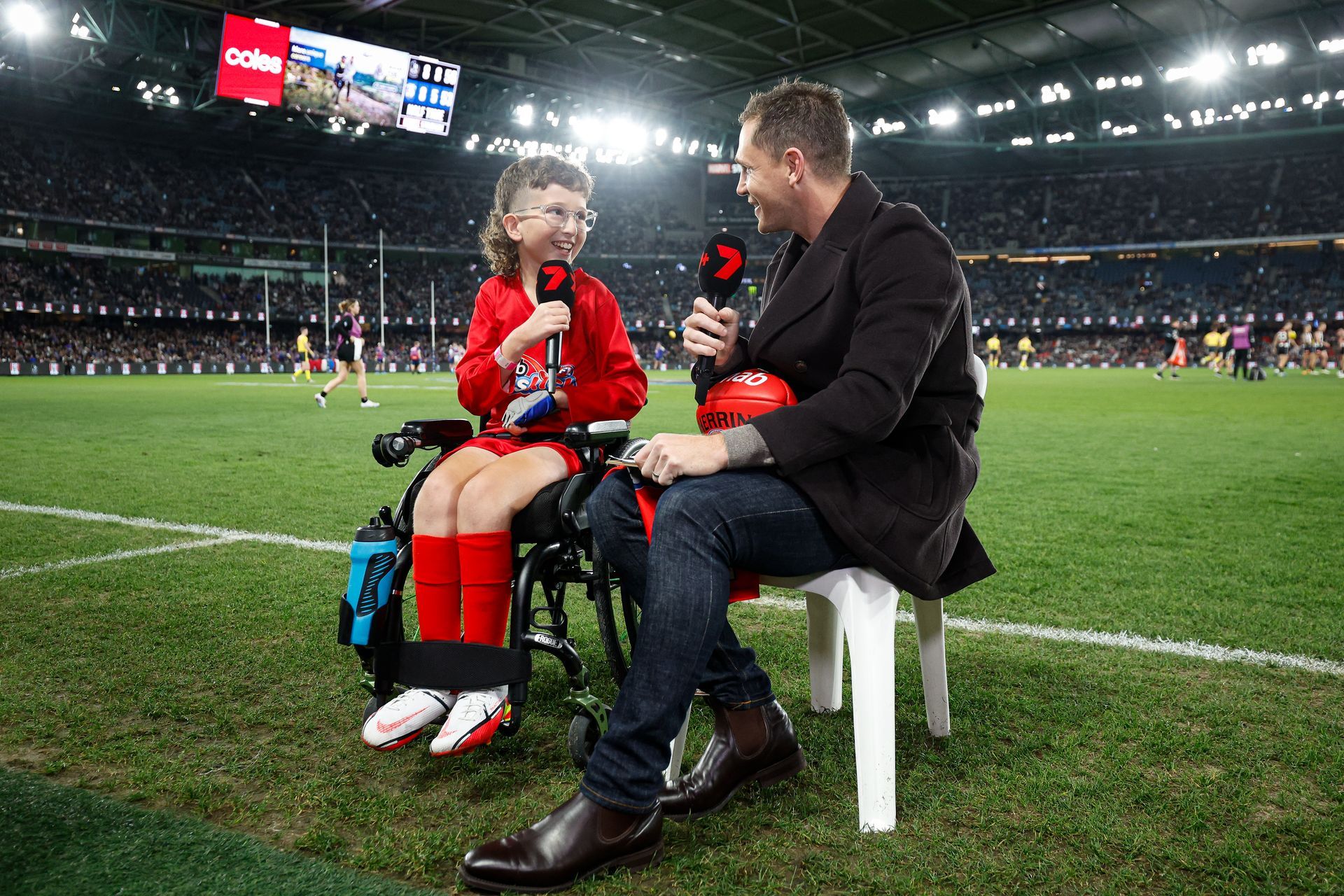 Recipient Alex wears an Auskick uniform as he is being interviewed on the sidelines of the MCG by Joel Selwood. Alex is a wheelchair user.
