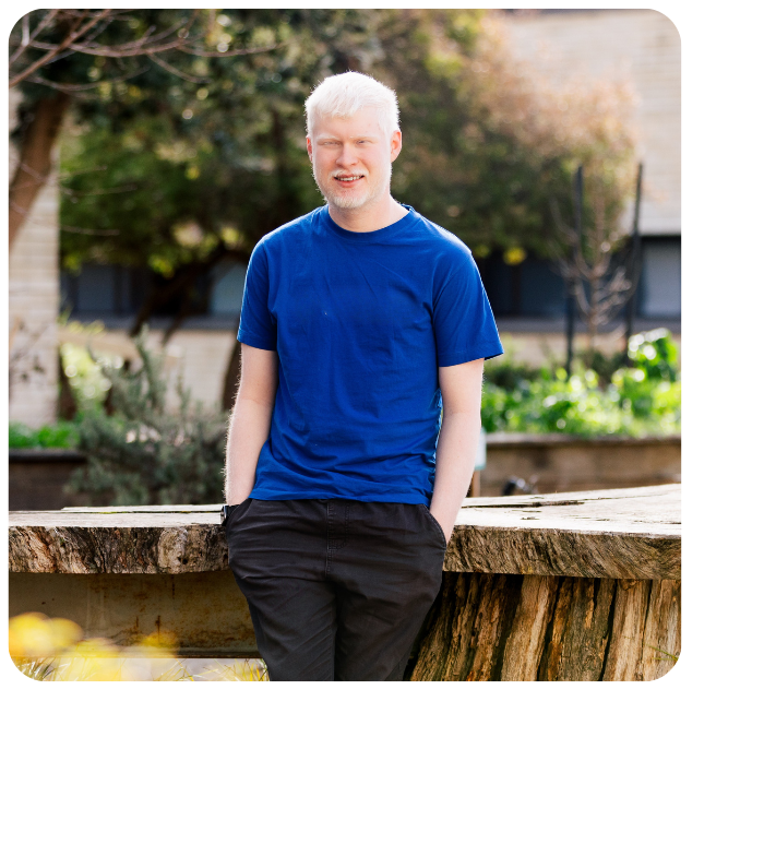 Jacob stands, hands in pockets, outside against a wooden table, smiling to camera in a blue shirt. 