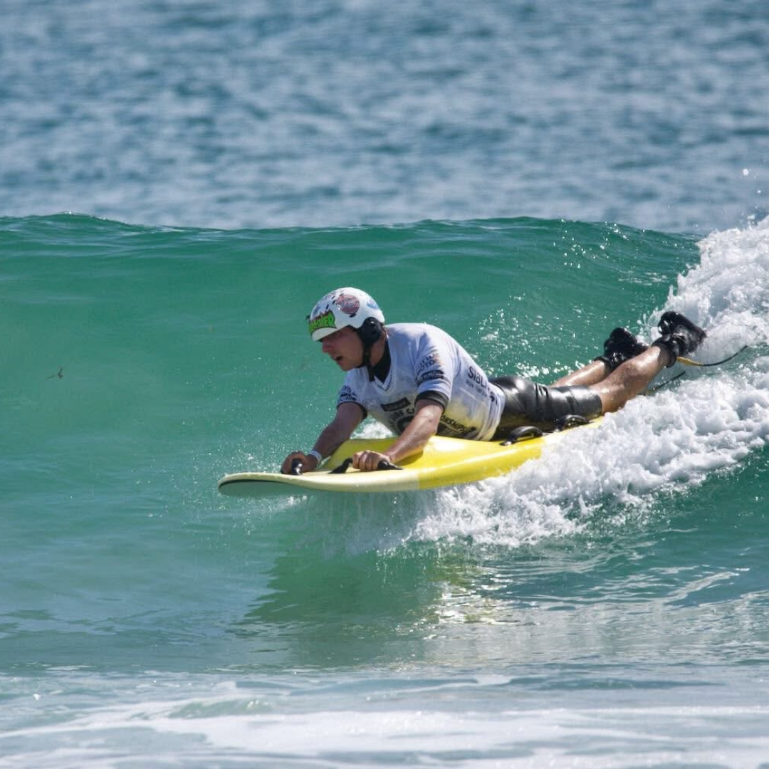 Bailey wears a helmet, wetsuit and white rash vest, laying on a yellow surfboard riding across the face of a wave.