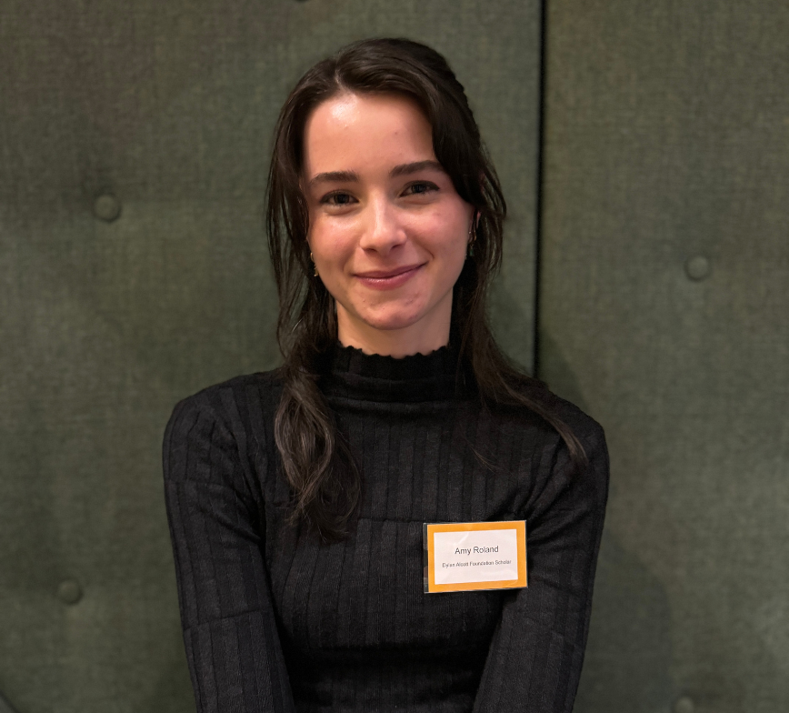 Close up shot of Amy sitting on a dark green couch smiling to camera. She has long black hair and wears a black long sleeve. A white and yellow name badge is pinned to her shirt.