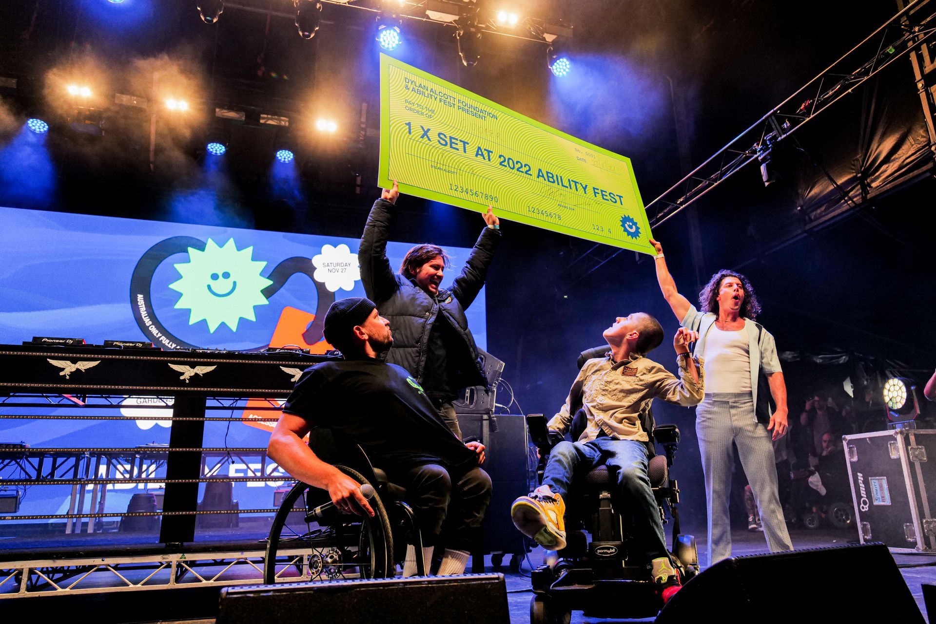 Dylan and recipient Cooper, on stage at Ability Fest looking at Peaking Duck holding a giant cheque above their heads. 