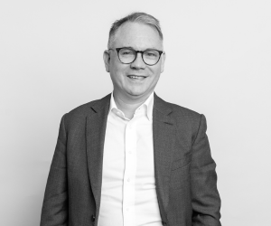 Black and white studio photo of Colin. He wears a suit and glasses.