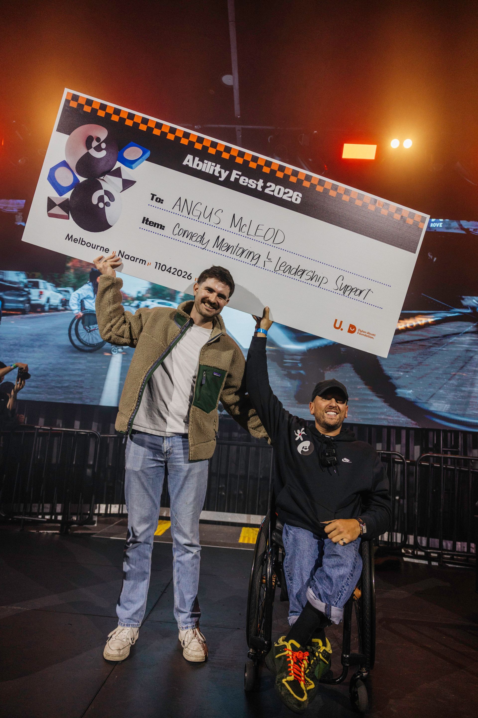 Angus (left) stands next to Dylan (right) who is a wheelchair user. They hold a giant cheque above their heads on the Ability Fest main stage.