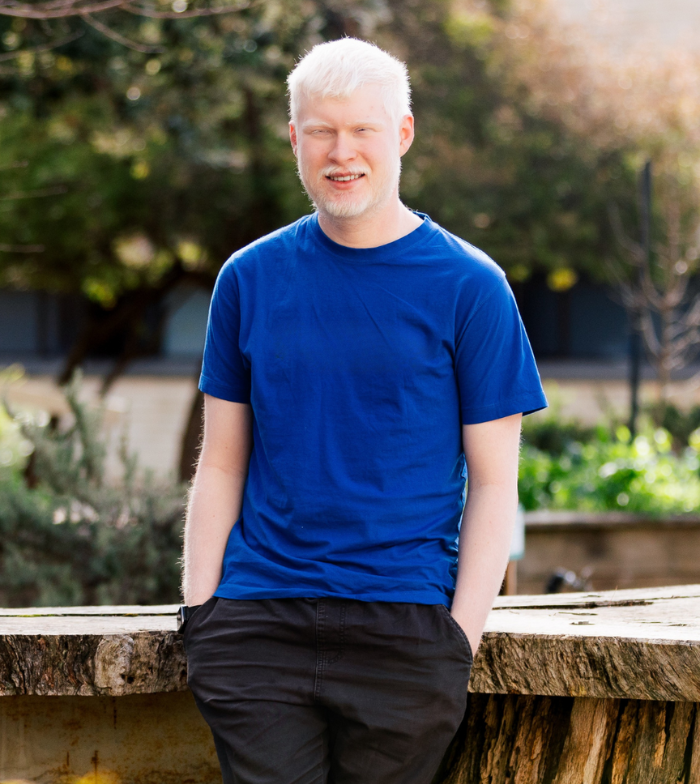 Jacob stands, hands in pockets, outside against a wooden table, smiling to camera in a blue shirt. 