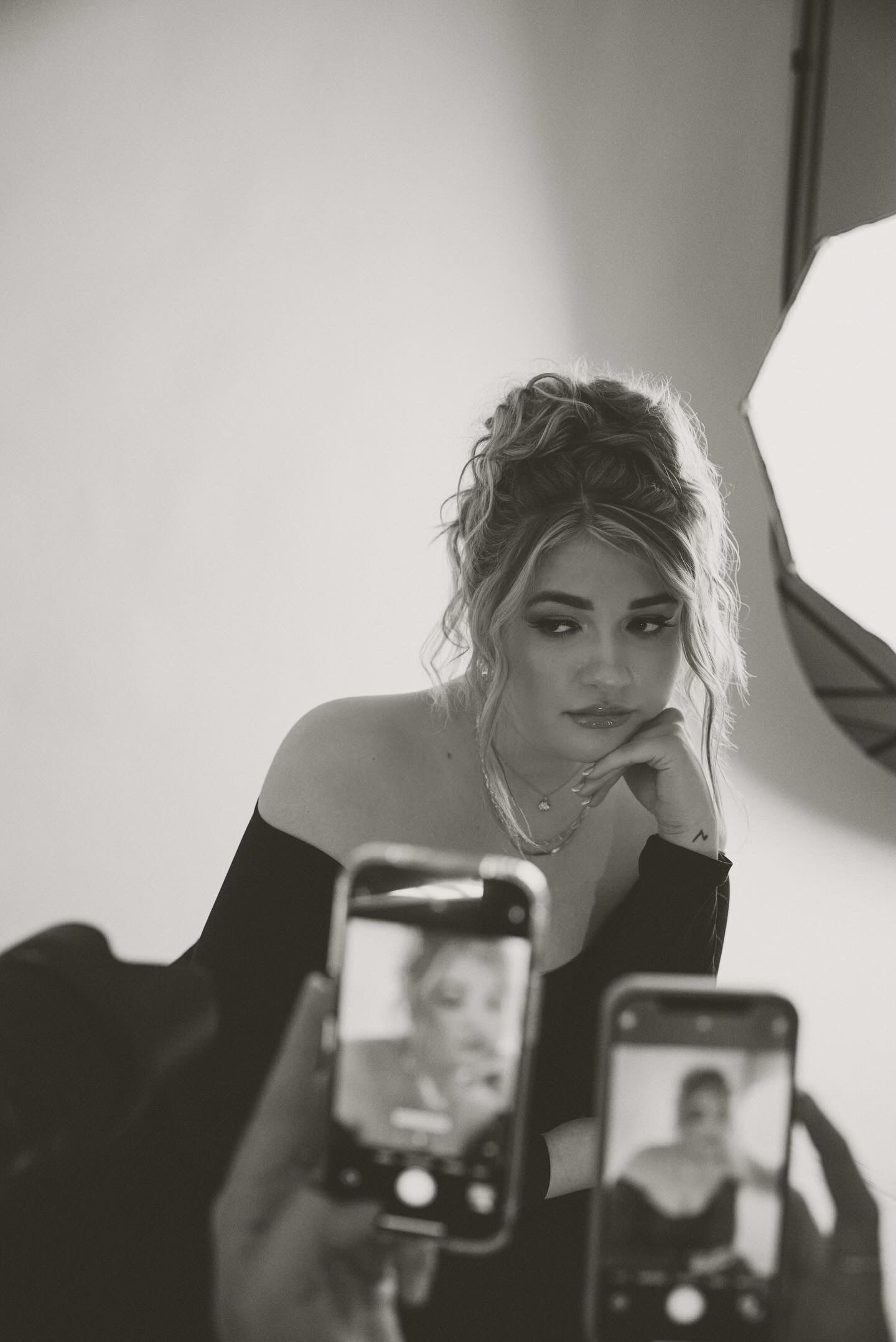 Black and white photo of Liv posing with her left hand resting under her chin during a photo shoot. Two phones are held in the foreground taking photos.