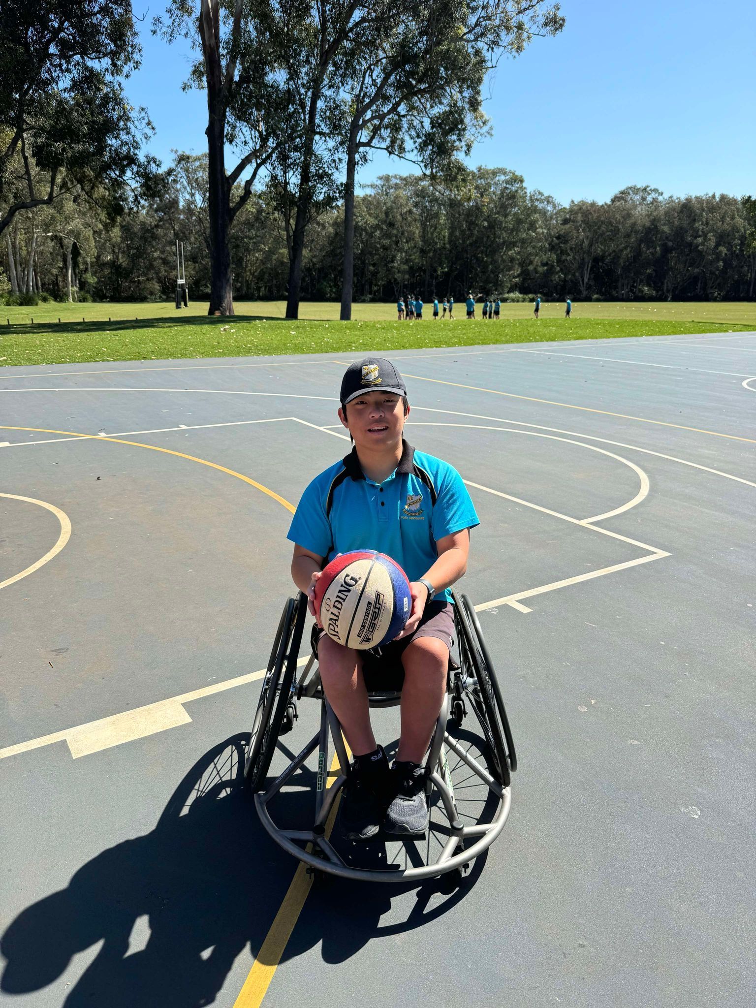 Max smiles to camera and holds a basketball court whilst in his sports wheelchair. He is outside on a sunny day at an outdoor basketball court.
