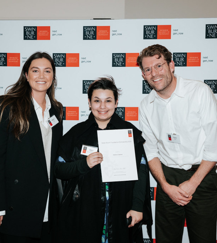 Elektra pictured holding a scholarship certificate in front of a Swinburne Uni media all. She is flanked by DAF staff members Stef (left) and Charlie (right) - all smiling to camera. Elektra holds a walking cane in one hand. 