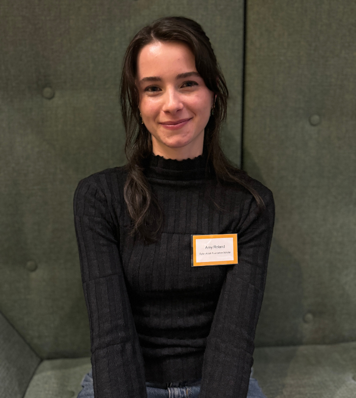Close up shot of Amy sitting on a dark green couch smiling to camera. She has long black hair and wears a black long sleeve. A white and yellow name badge is pinned to her shirt. 