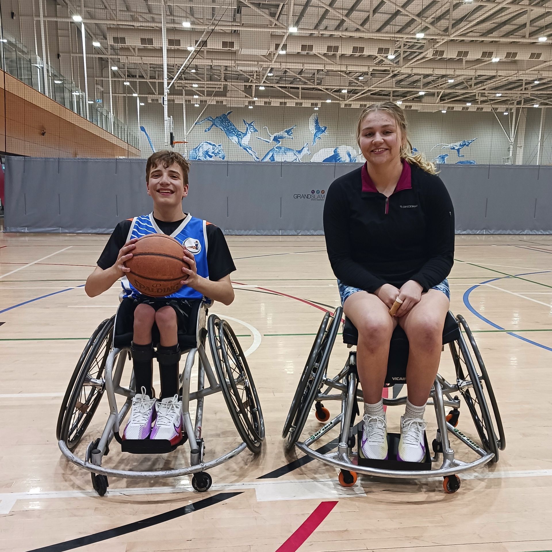 Alex (left) and Isobella (right) seated in their basketball wheelchairs, smiling at the camera. Alex holds a basketball at his chest and both are wearing active wear. The shot is taken at an indoor court.