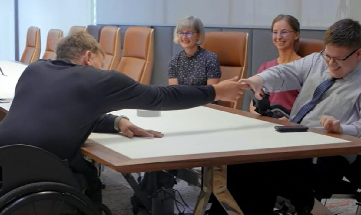 Dylan (left) in a board room shaking hands with recipient Cameron (right) across a table.