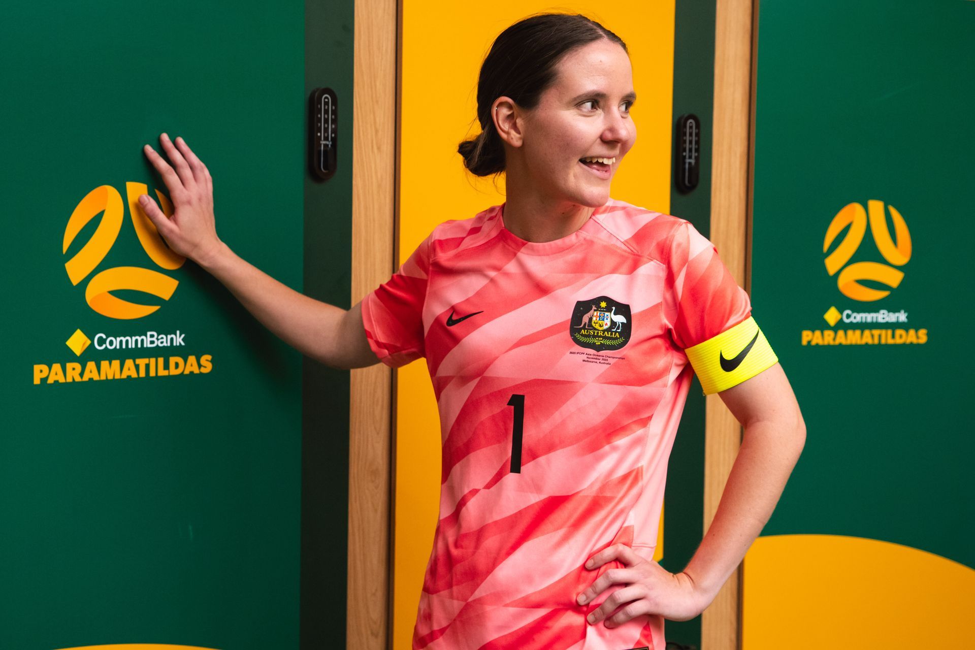 Carly rests her left hand on a locker that reads ParaMatildas, whilst looking behind over her shoulder smiling. She wears a goalie ParaMatildas football top with a yellow captain arm band.