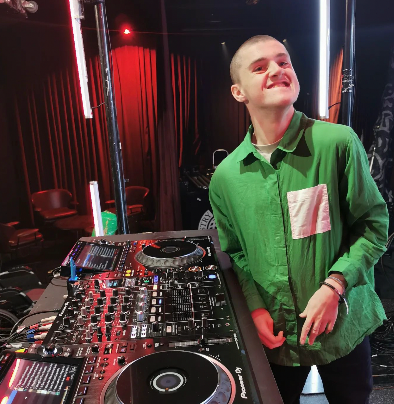 Cooper stands next to DJ decks smiling to camera.