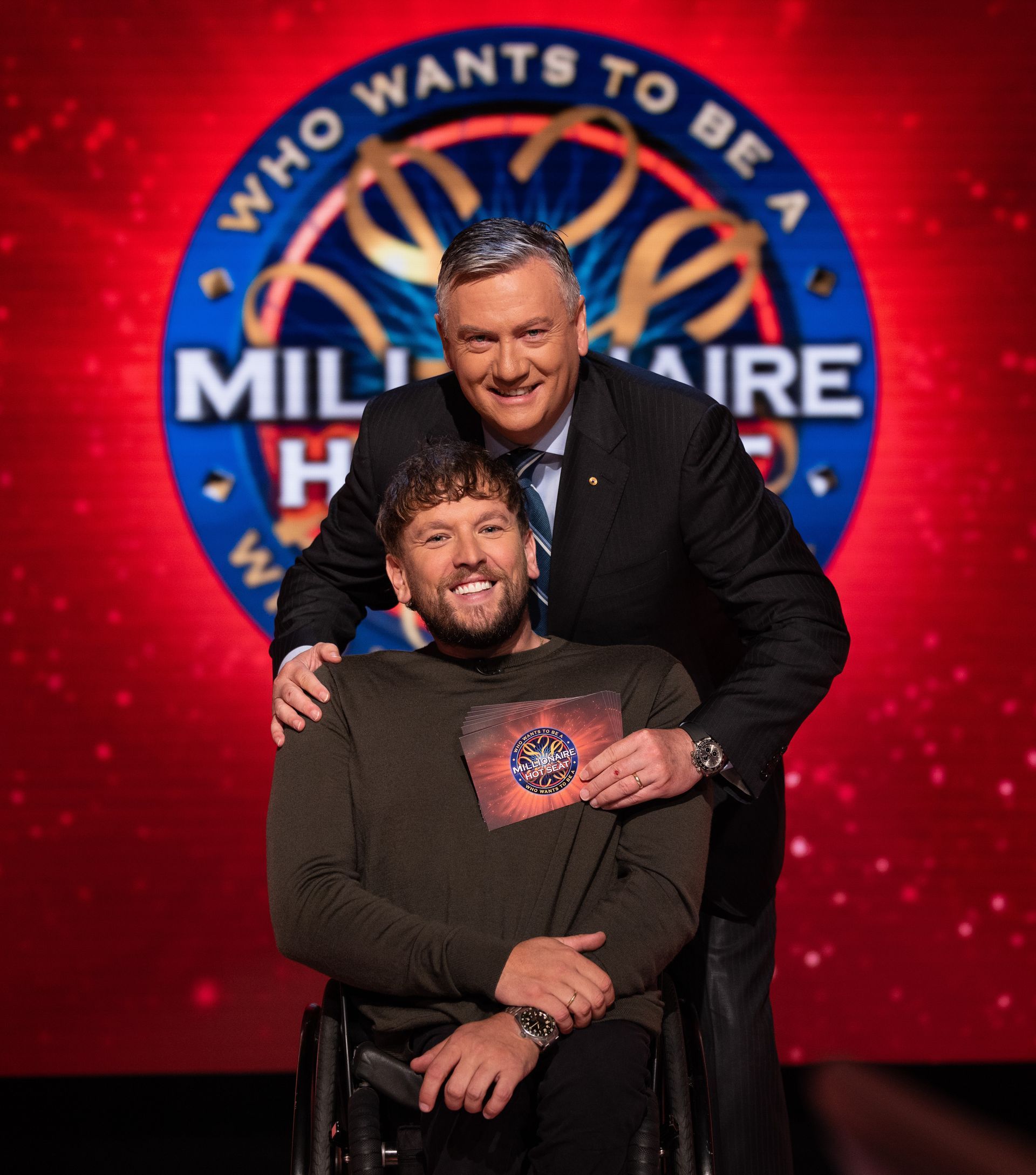 Dylan middle with Eddie McGuire behind smiling to camera. The the red background displays the blue Millionaire Hot Seat logo. 