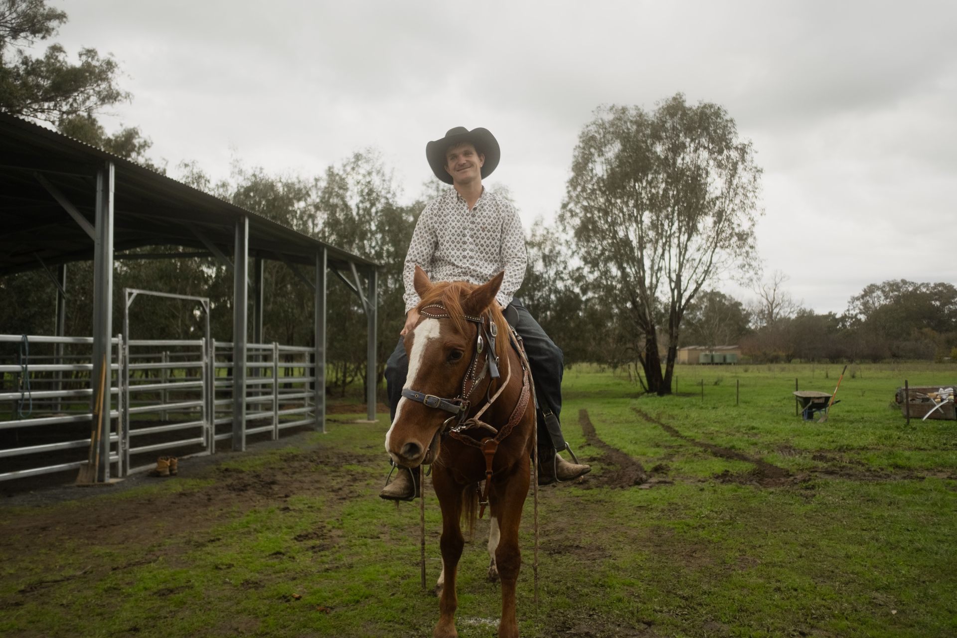 Brodie on a farm atop a horse, facing the camera and smiling. He wears jeans cowboy boots and hat.