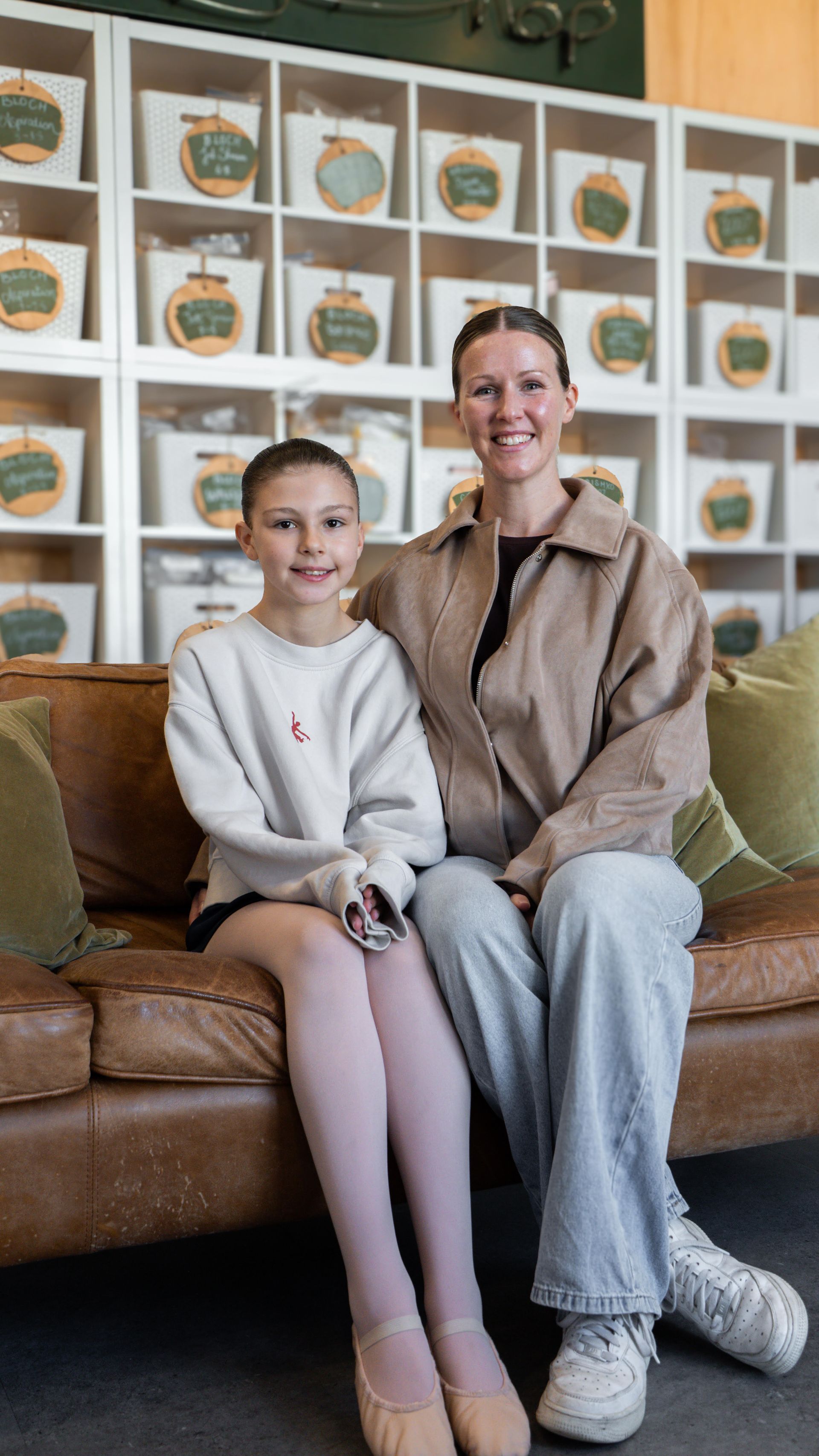 Delilah (left) with mum, Lisa(right), sit side by side on a brown leather couch. The background shows a wall of baskets with ballet shoe tags. Delilah wears ballet tights and shoes.