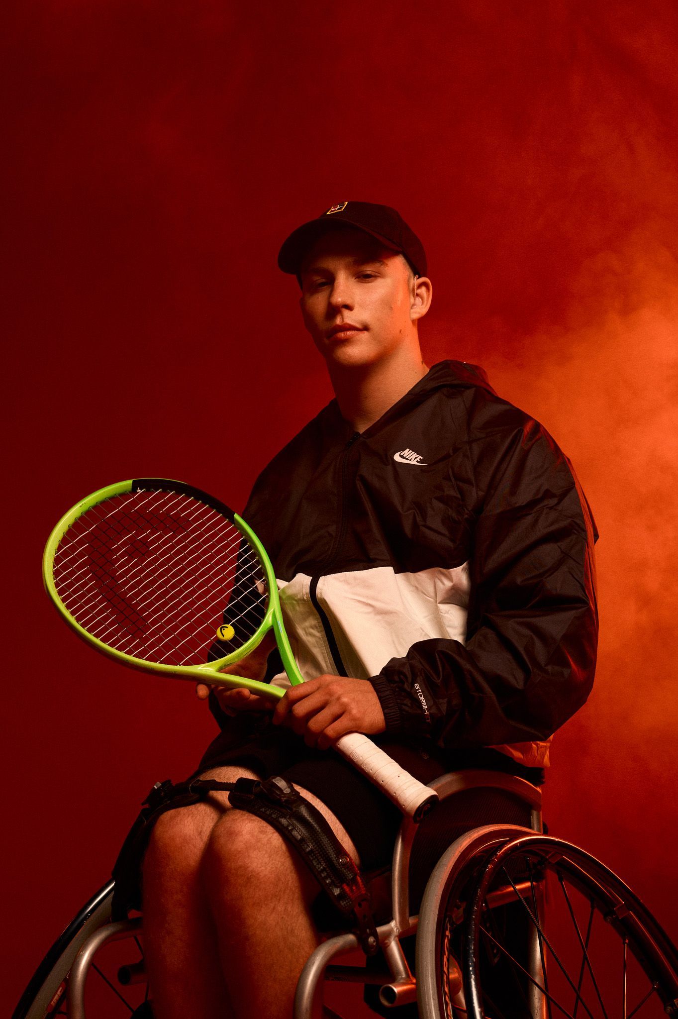 Studio image of Ben, he sits in a tennis wheelchair holding a tennis racquet, wearing Nike kit. He is in front of a red background and smoke in the air.