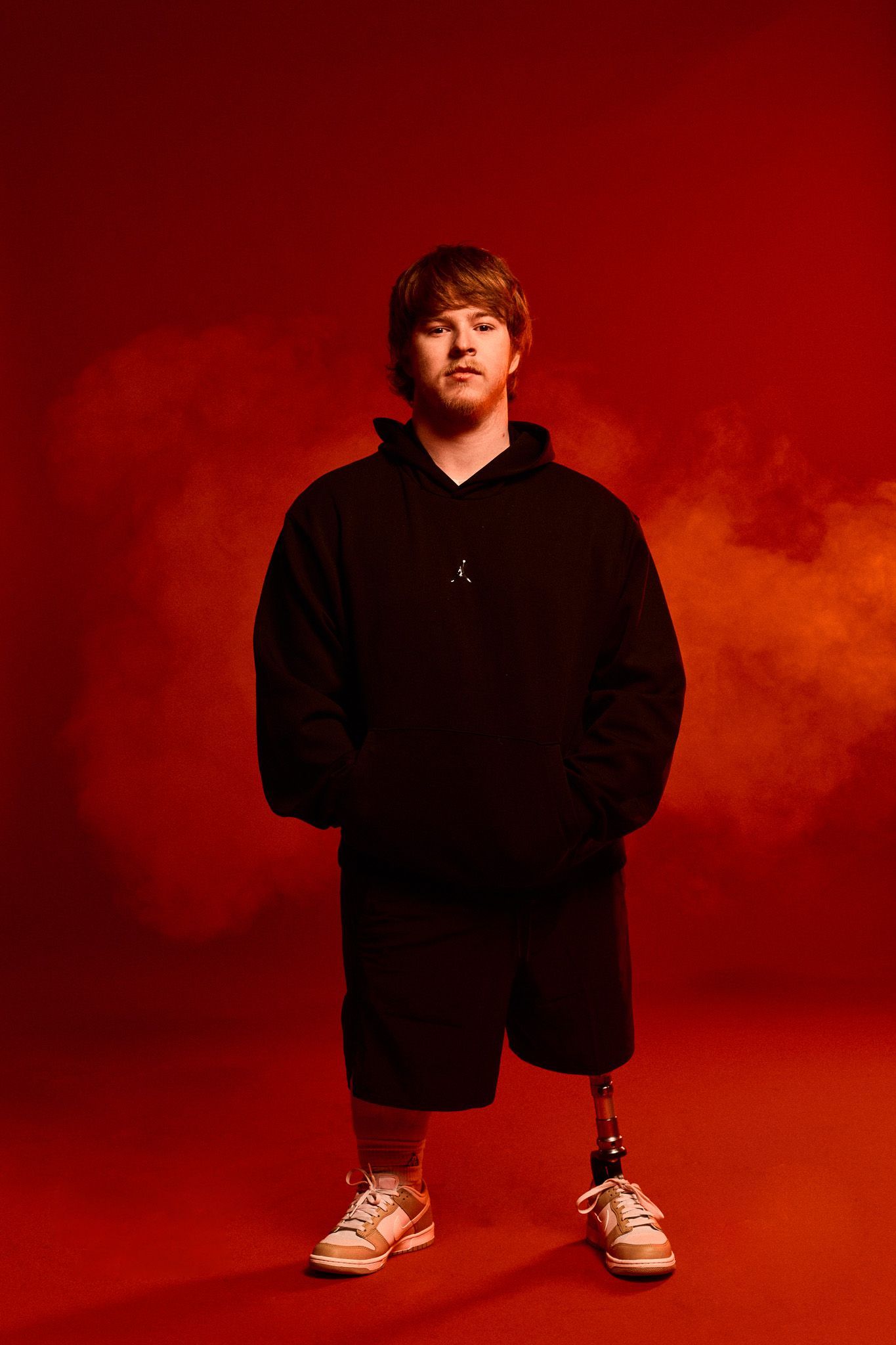 Studio image of Brayden, arms in his pockets, wearing Nike kit. He stands in front of a red background and smoke in the air. Brayden wears a prosthetic left leg.