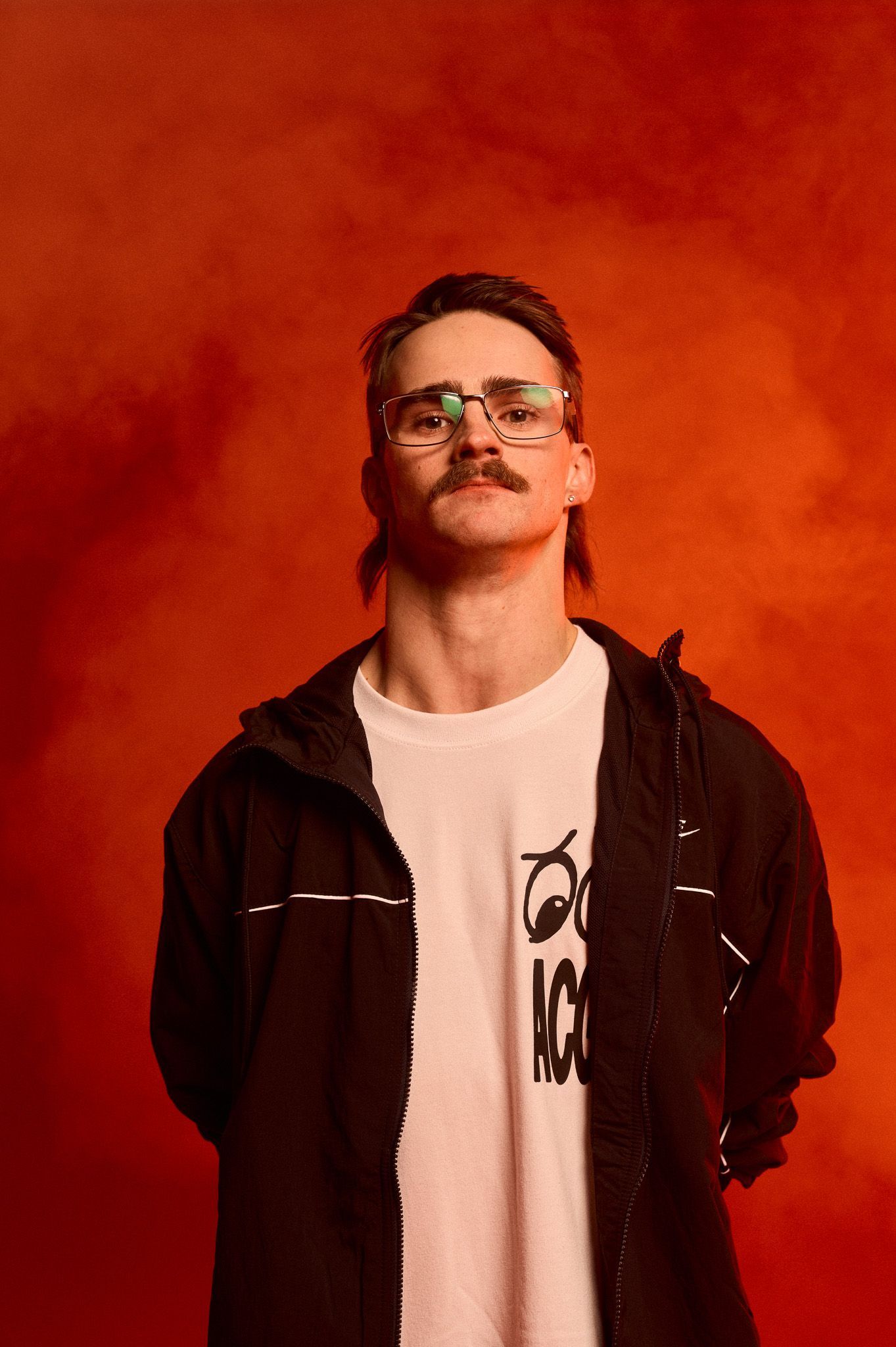 Studio image of Glen, arms behind his back, chin tilted up, wearing Nike kit. He stands in front of a red background and smoke in the air.