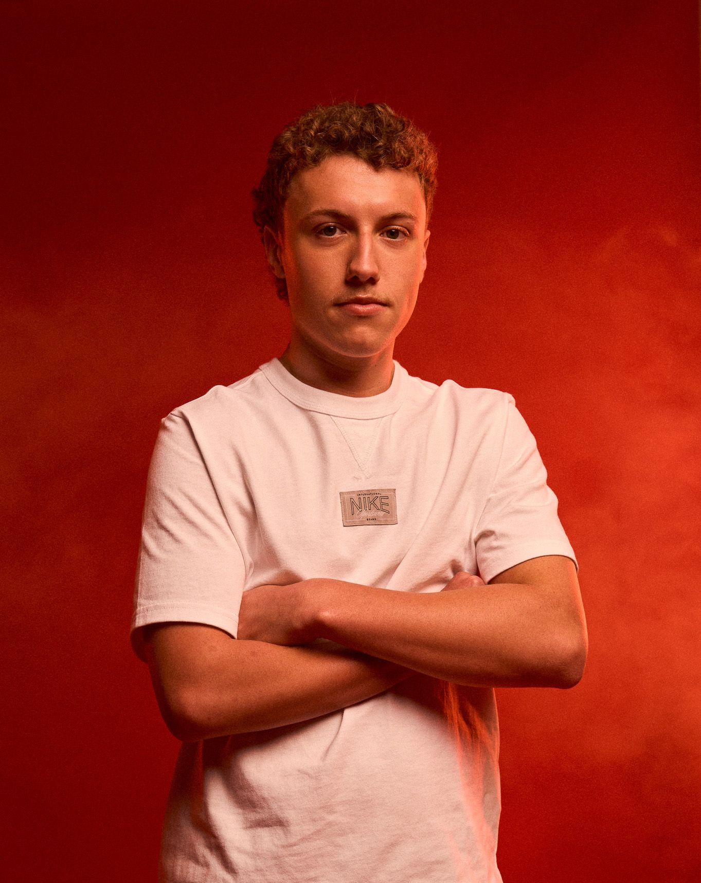 Studio image of Matthew, arms crossed, wearing Nike kit. He stands in front of a red background and smoke in the air.