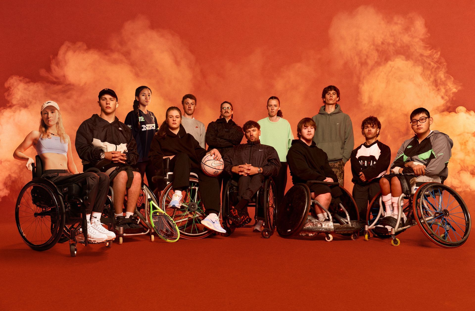 Group photo of eleven athletes crowded around Dylan, all with varying disability. All individuals wear Nike kit. Two female and four males in the front row are wheelchair users. The studio backdrop is red with smoke lingering in the air behind athletes.
