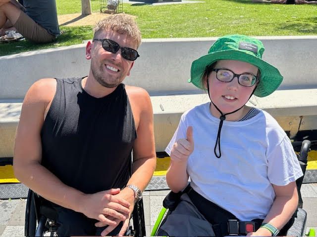 Dylan (left) pictured with recipient Hugh (right) smiling to camera. They are outside in the sunshine with both individuals being wheelchair users.