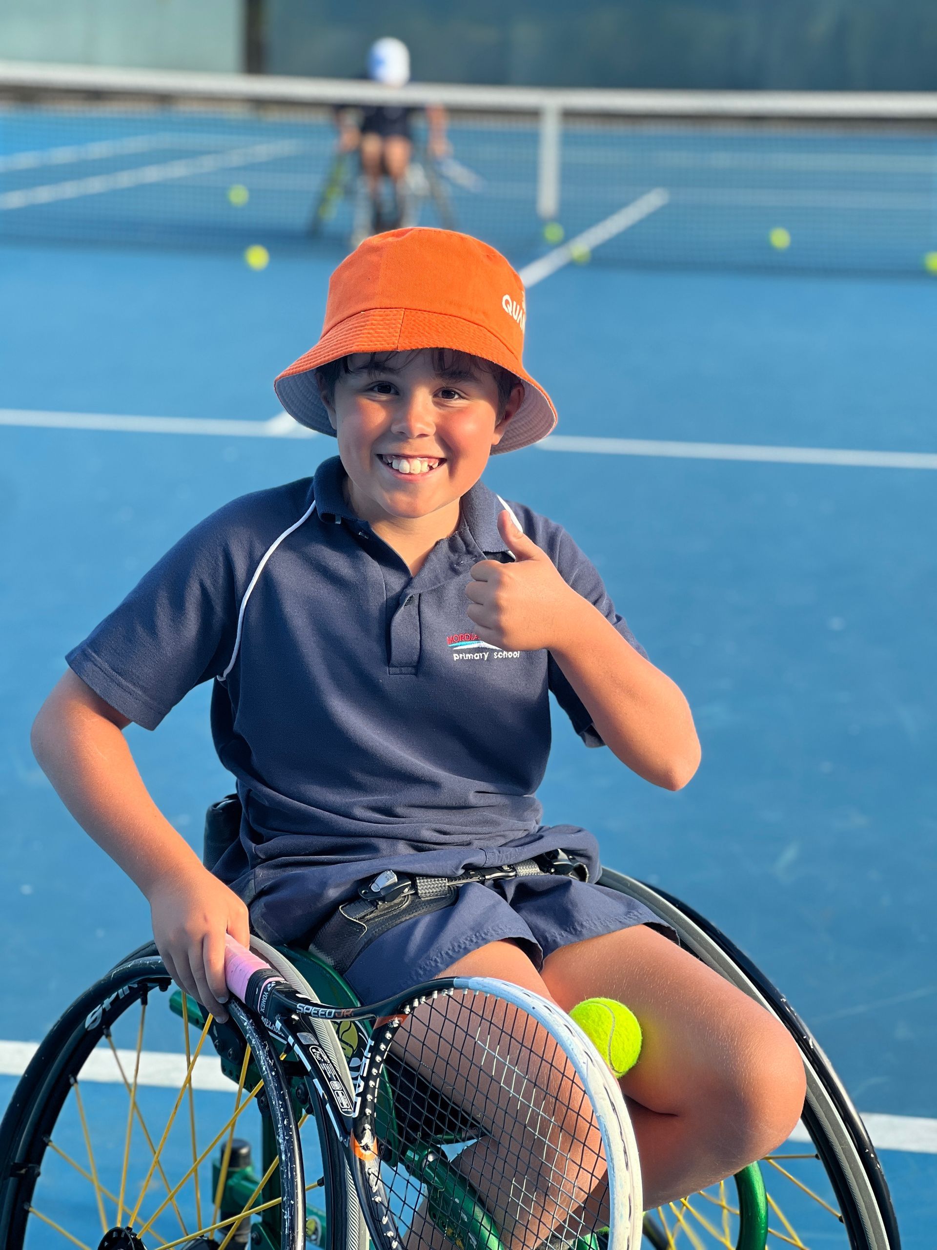 Sonny gives a thumbs up to the camera in his tennis wheelchair. The background shows a blue tennis court and tennis balls on the ground.