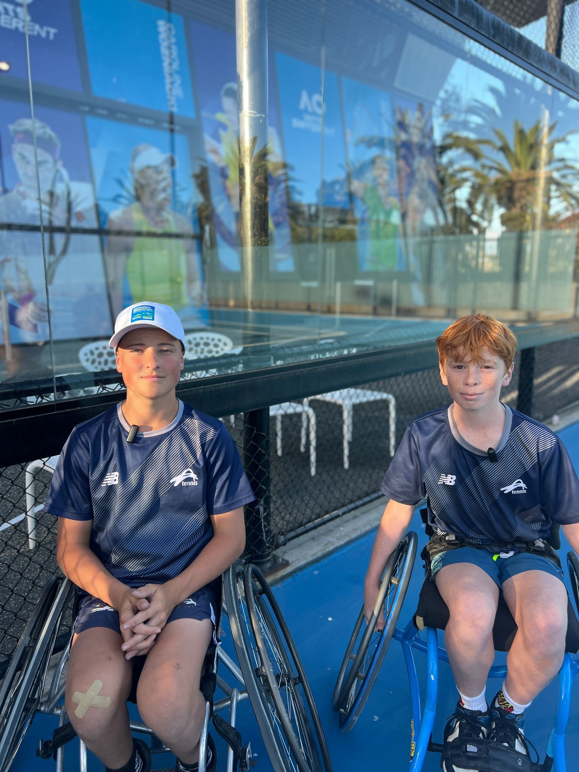Gillie (left) and Arlo (right) smile to camera on an outdoor blue tennis court in their tennis specific wheelchairs.