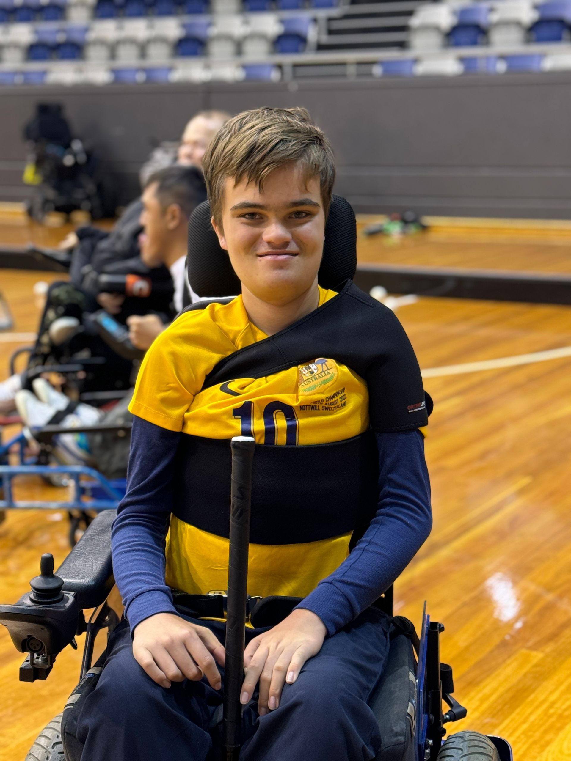 Daniel wears a yellow jersey with the number 10 and holds a hockey stick in both hands. He's at an indoor basketball court and in his new DAF power chair. 