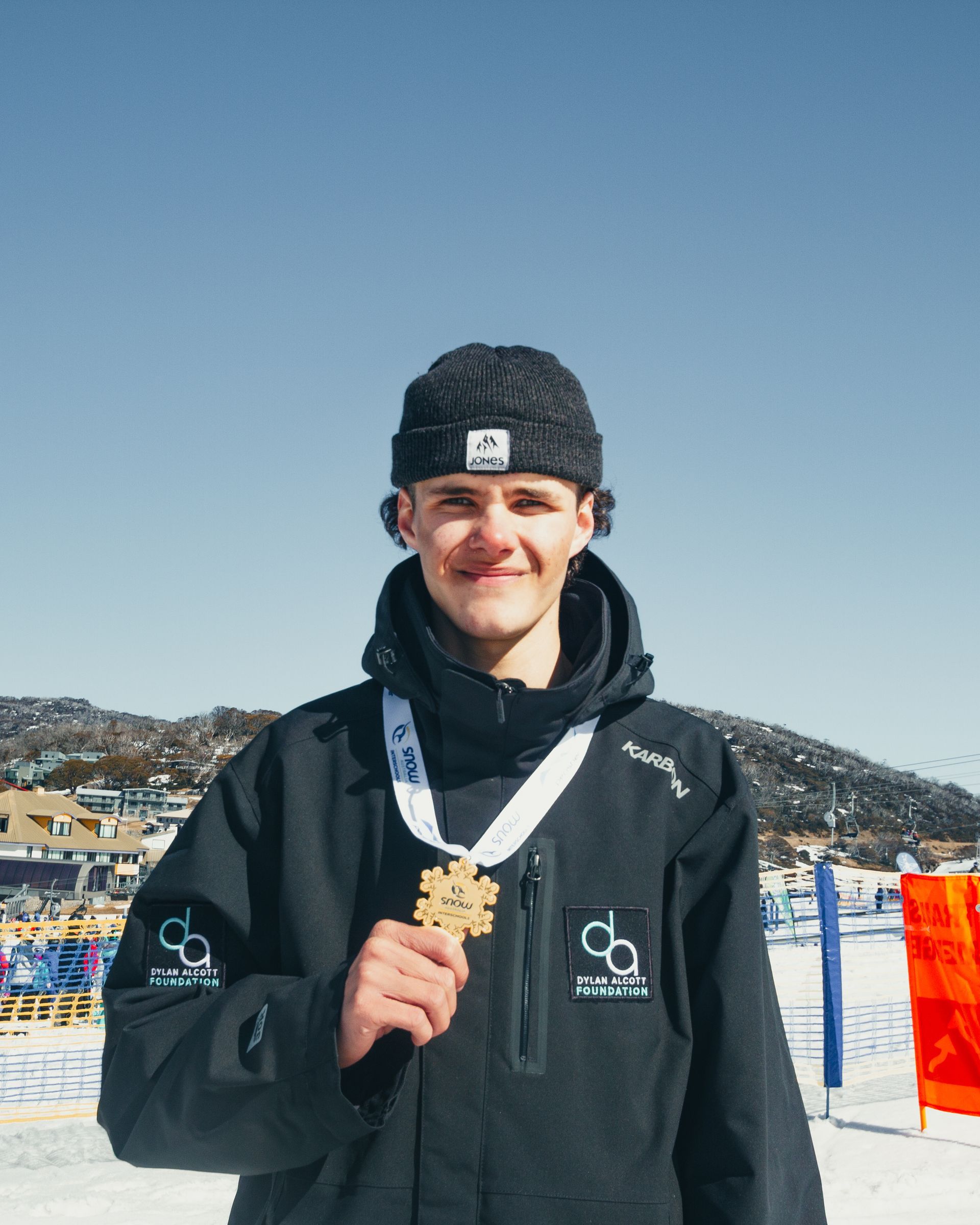 Recipient Lachlan wears a snow jacket and beanie with a first place medal around his neck. Snow mountains are in the background.
