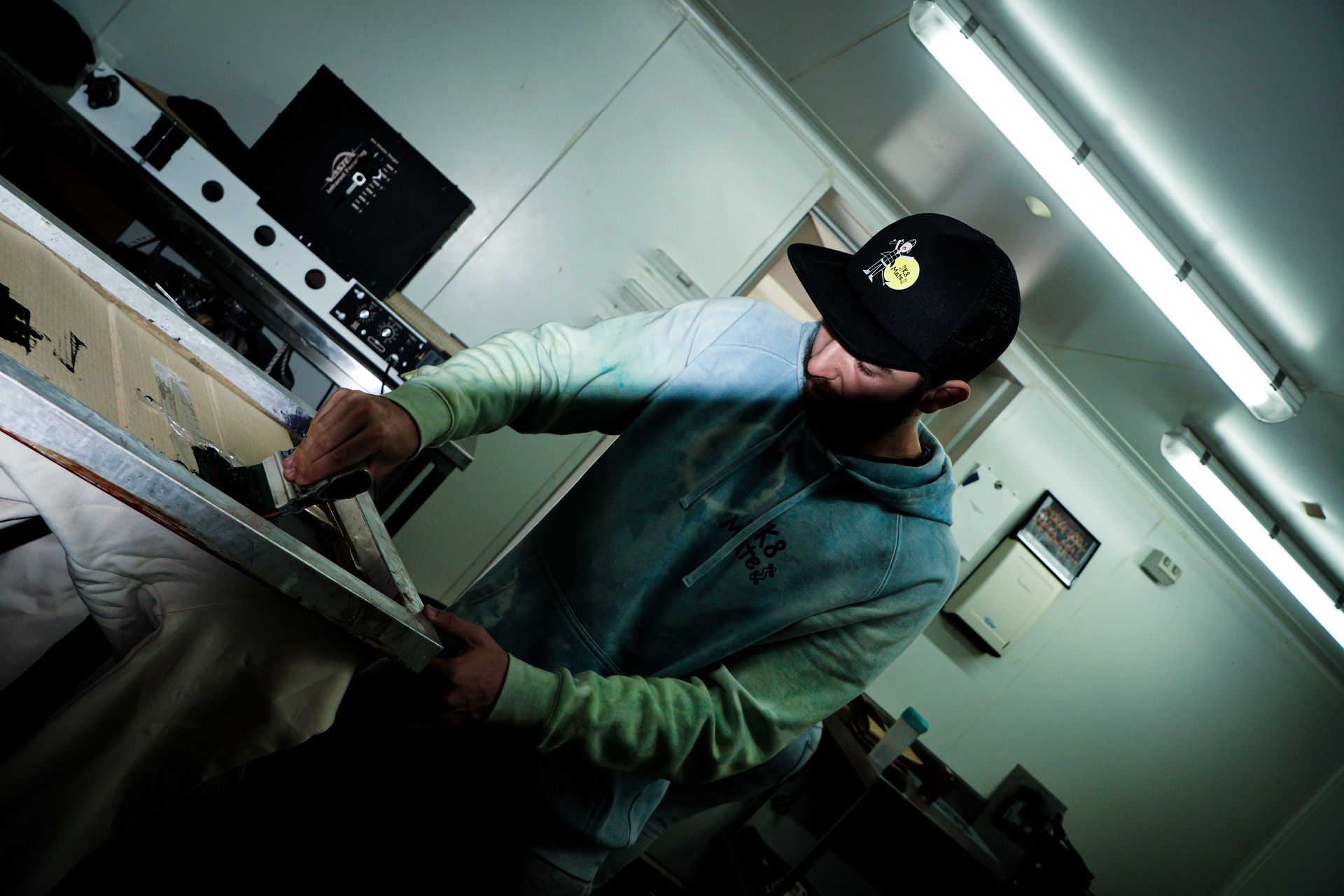 Seb wears a hat and tie dyed jumper as he glides black ink across a screen printer onto a jumper. The background shows a screen printing work room with benches.