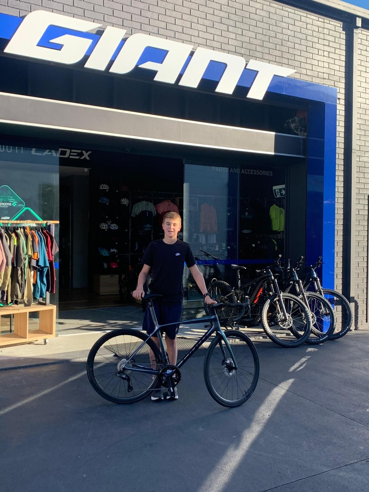 Jack poses out the front of a Giant store with his black and dark grey bike. The store front is blue with white capital letters at the top reading “GIANT”.