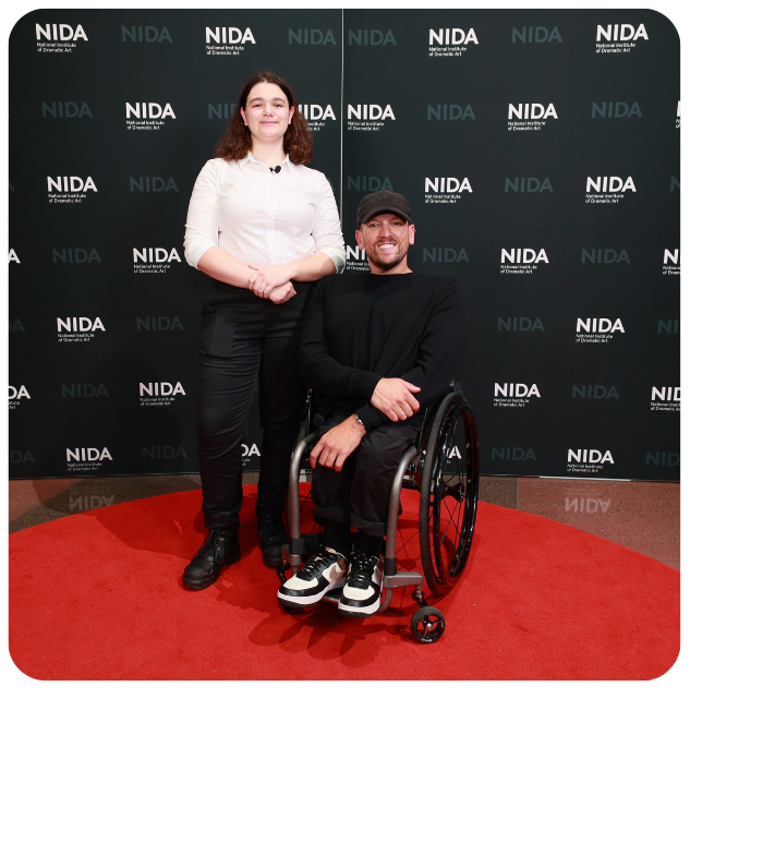 Kaia (left) stands next to Dylan (right) who is a wheelchair user. They smile to camera on red carpet with a black and white NIDA media wall behind them.