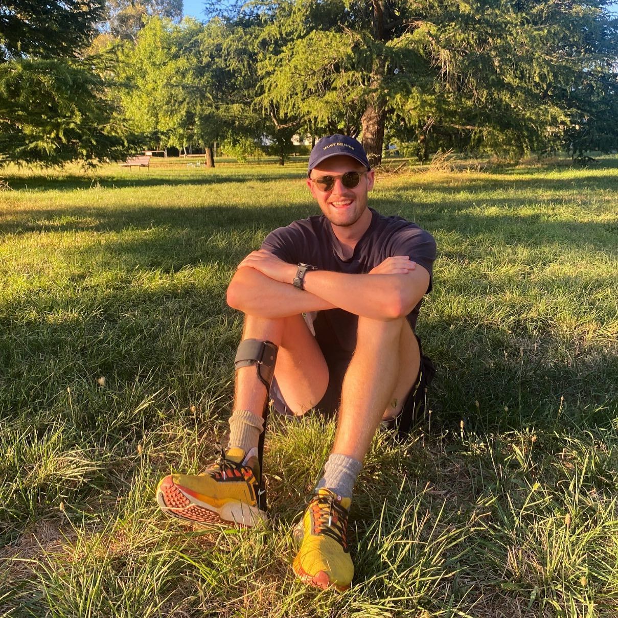 Nick sits in a park on green grass with trees in the background. Smiling at the camera, with arms crossed over his knees. He wears exercise gear, plus a custom made Ankle Foot Orthotic.