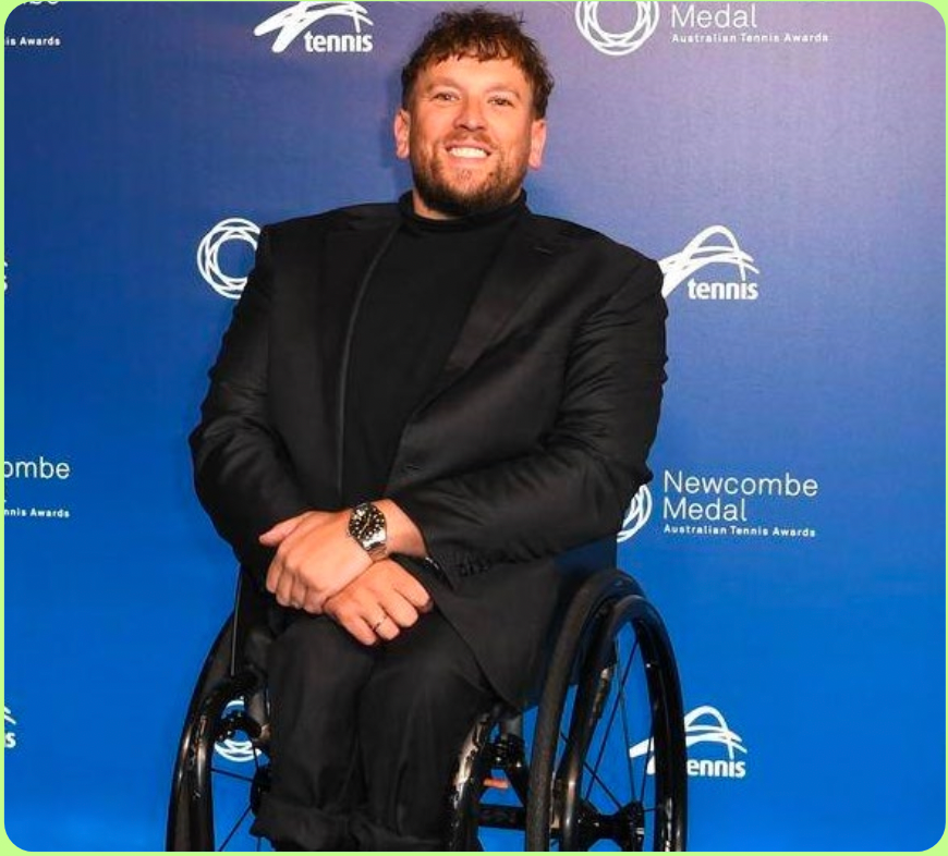 Man in a black suit seated in a wheelchair at a blue event backdrop with tennis logos