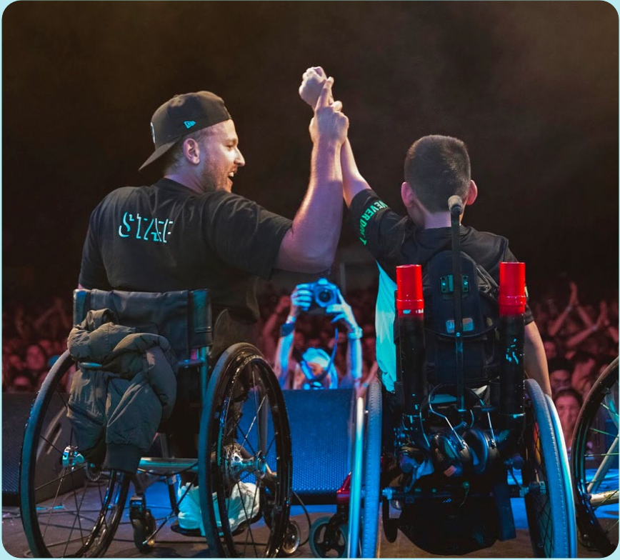 Shot from behind of recipient Jin (right) and Dylan (left) holding hands raised on stage at Ability Fest. Both are wheelchair users.